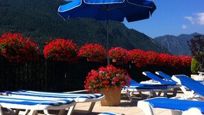 Un espace piscine avec des chaises longues à rayures bleues et blanches, un grand parasol bleu et des fleurs rouges en arrière-plan.