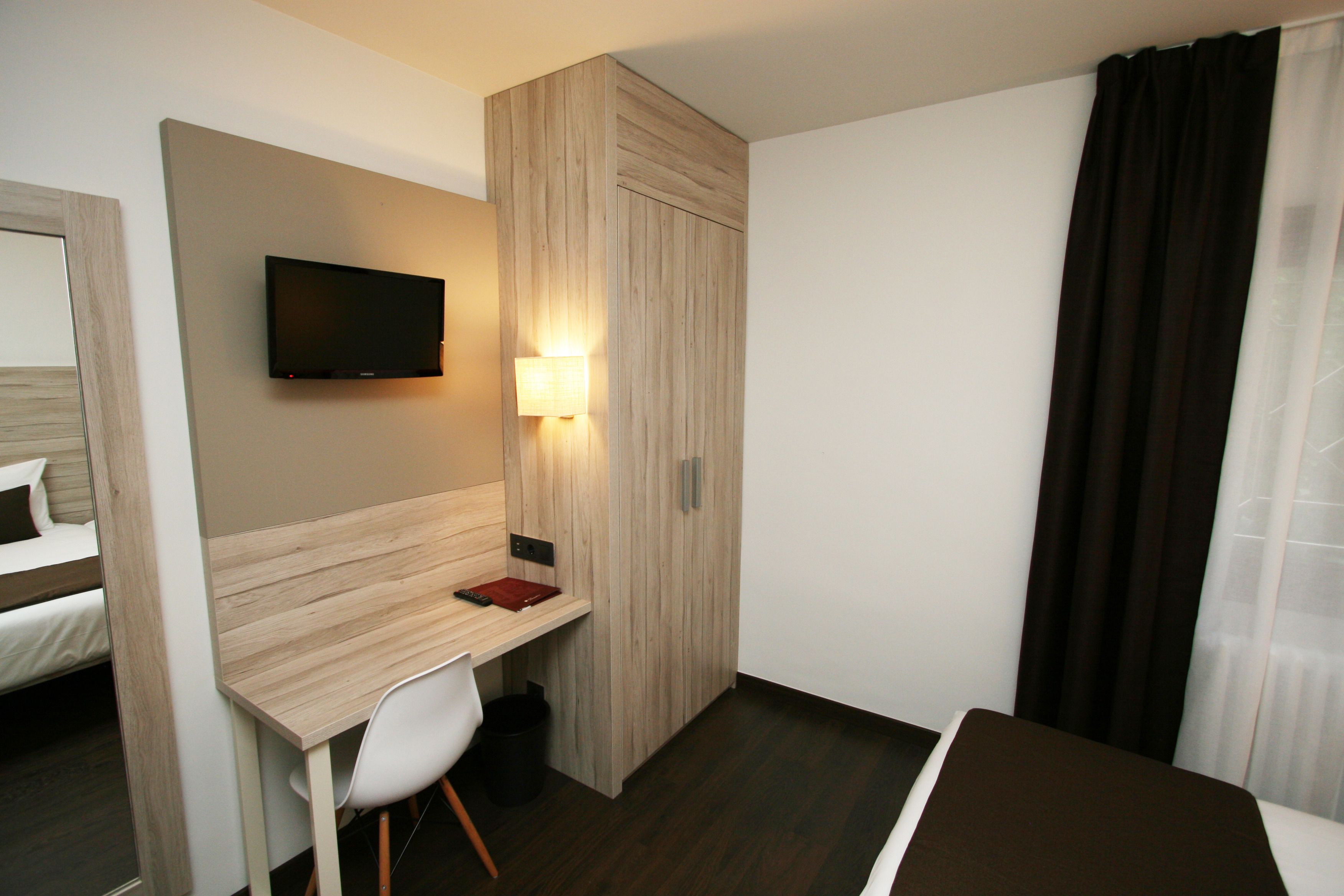 Quarto de hotel com cama, escrivaninha, cadeira, televisão na parede e janela com cortinas.