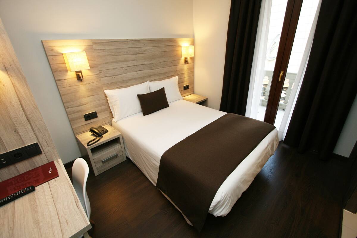 Ein Hotelzimmer mit einem großen Bett, zwei Nachttischen mit Lampen, einem Schreibtisch und einem Stuhl sowie einem Blick ins Freie durch ein Fenster.
