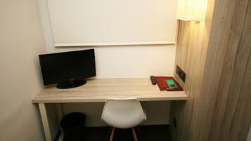 Un pequeño espacio de trabajo con un escritorio, una silla, un monitor de computadora, un control remoto y una lámpara.