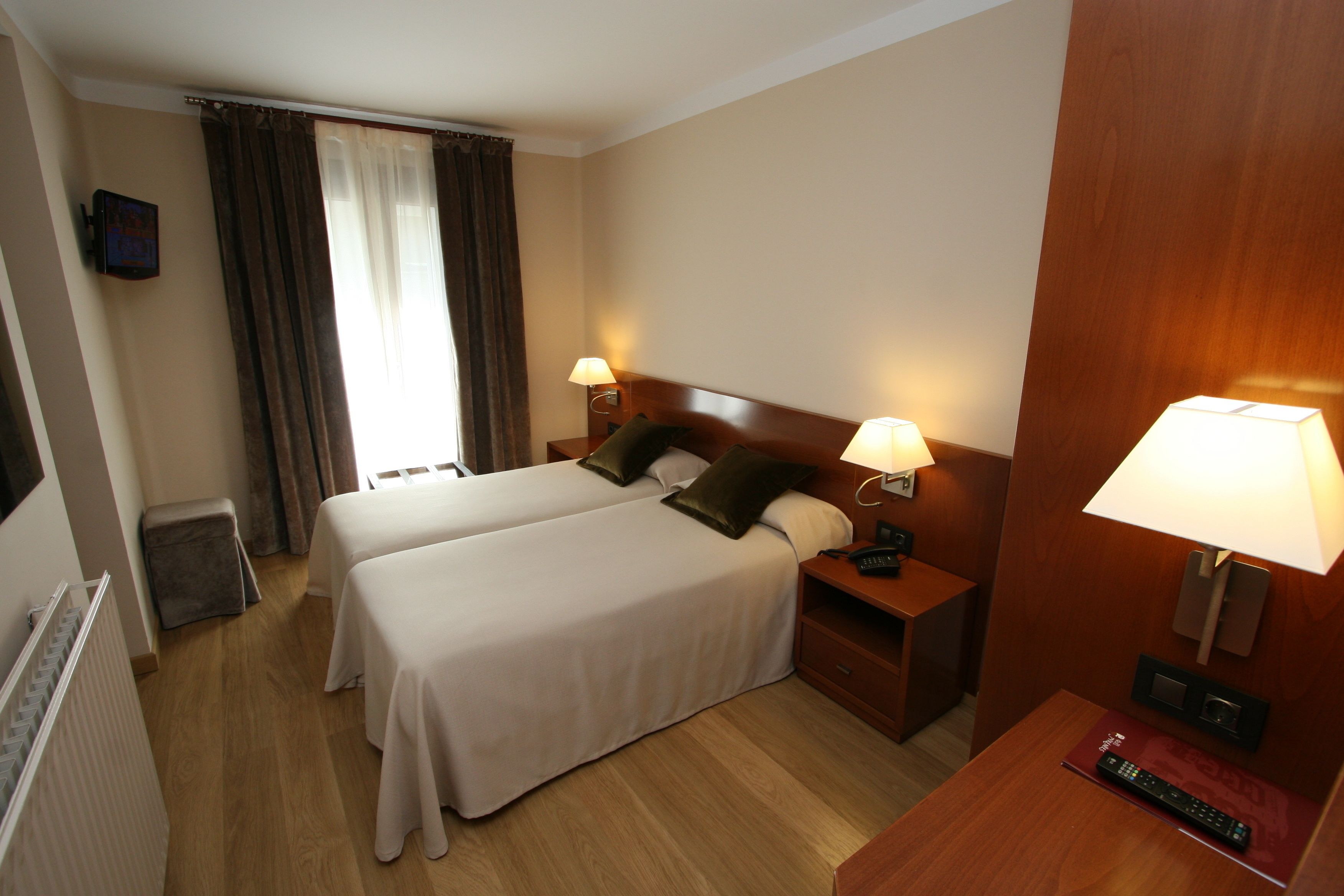 Habitación de hotel con dos camas, cabecero de madera, lámparas de noche, televisión y ventana con cortinas.