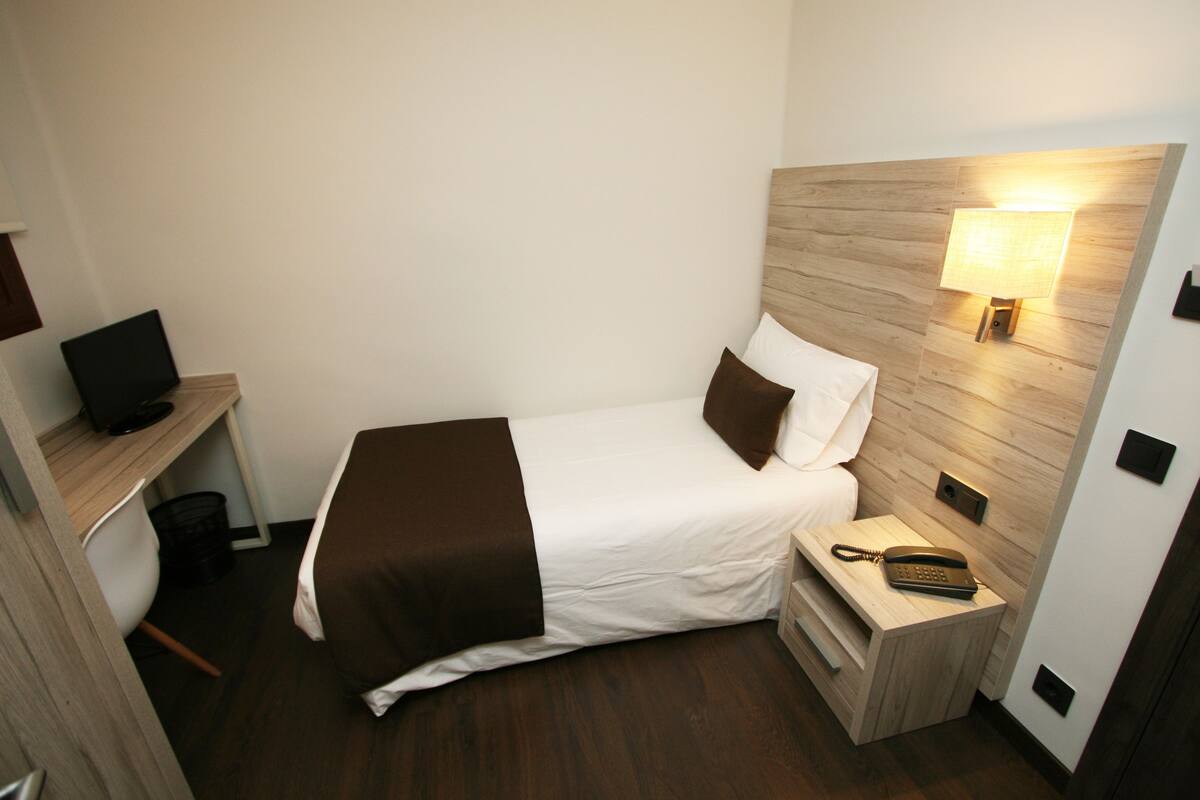 Ein kleines Hotelzimmer mit einem Bett, einem Nachttisch mit Lampe, einem Schreibtisch mit Computer und einem Telefon.