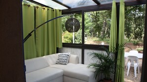 A corner living area with a white sofa, a patterned pillow, a hanging spherical light fixture, a potted plant, and a view of the outdoors through a window with green curtains. - Athénopolis (Grimaud)