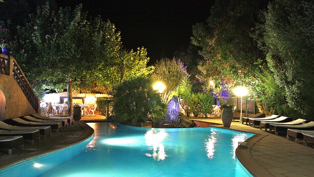 A swimming pool with lounge chairs and a waterfall feature, surrounded by trees and illuminated by night lighting. - Athénopolis (Grimaud)