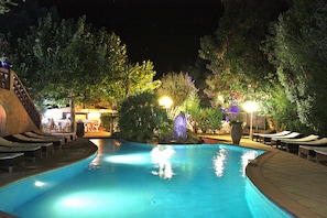 A swimming pool with lounge chairs and a waterfall feature, surrounded by trees and illuminated by night lighting. - Athénopolis (Grimaud)