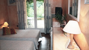 A bedroom with two beds, a desk with a lamp, a potted plant, and a view of the outdoors. - Athénopolis (Grimaud)