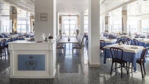 A spacious dining area with multiple tables and chairs, a central white counter, and large windows allowing natural light. - Capri (Jesolo)