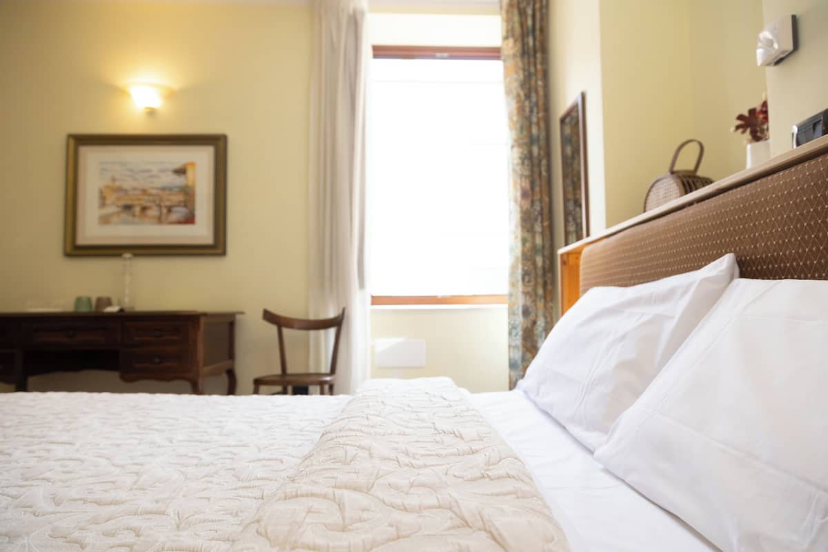 Habitación de hotel con una cama bien hecha, un escritorio con silla, un cuadro en la pared y una ventana con cortinas.
