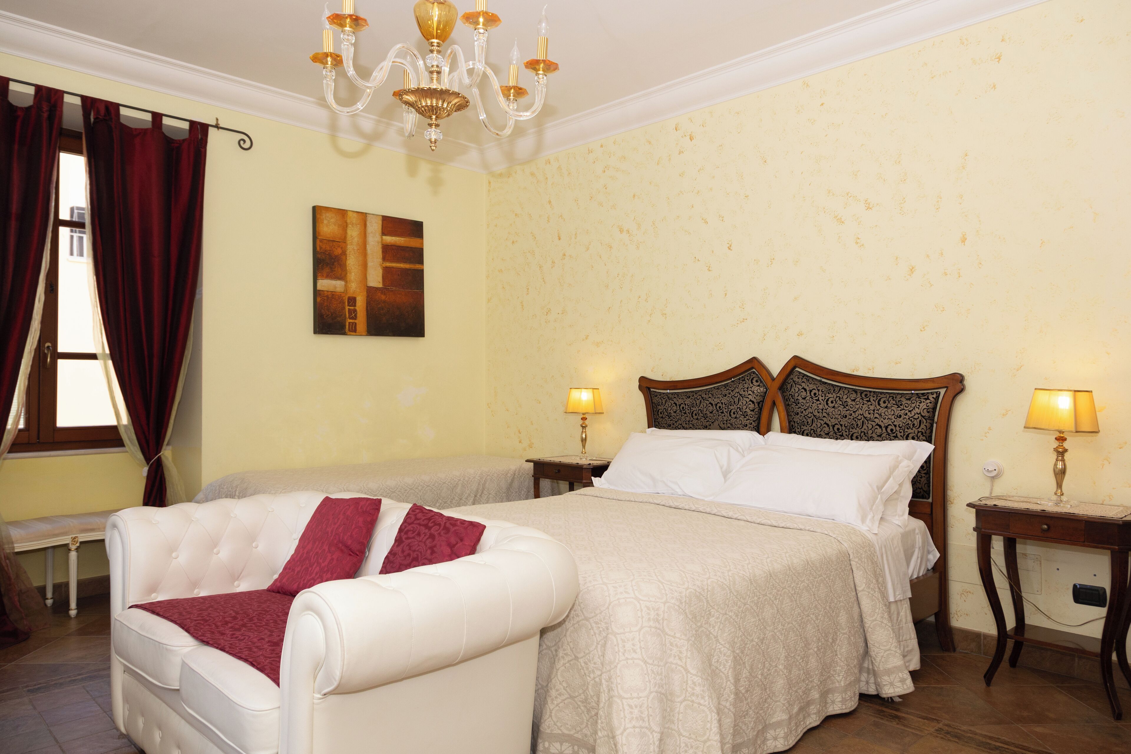 A hotel room with a large bed, a smaller sofa, two bedside tables with lamps, a chandelier, and a painting on the wall.