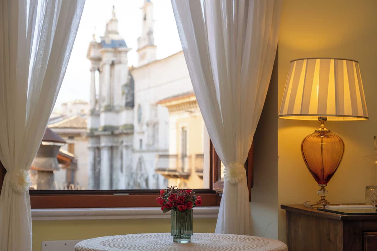 Una habitación con vista a un edificio histórico, un florero con flores sobre una mesa y una lámpara en una mesita de noche.