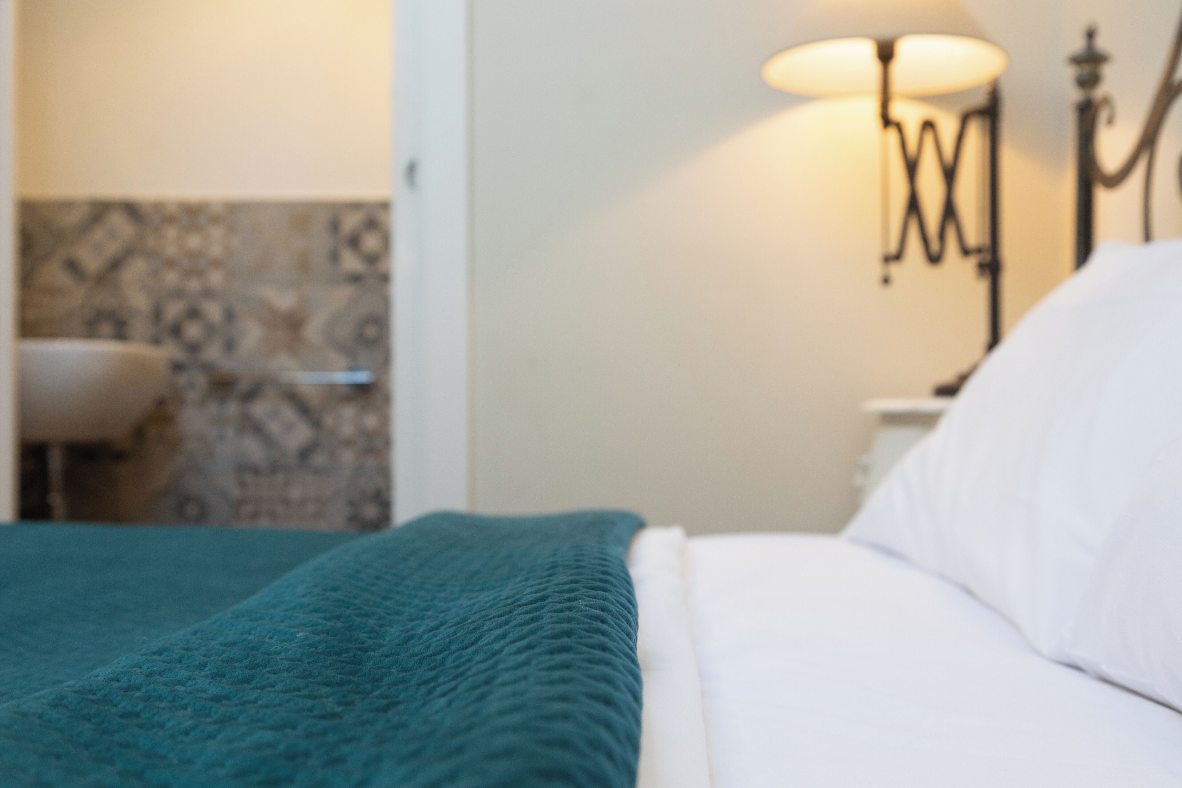 Una cama con una manta color aguamarina y almohadas blancas, una lámpara de pared, y un baño con lavamanos y baldosas estampadas.