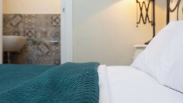 Una cama con una manta color aguamarina y almohadas blancas, una lámpara de pared, y un baño con lavamanos y baldosas estampadas.