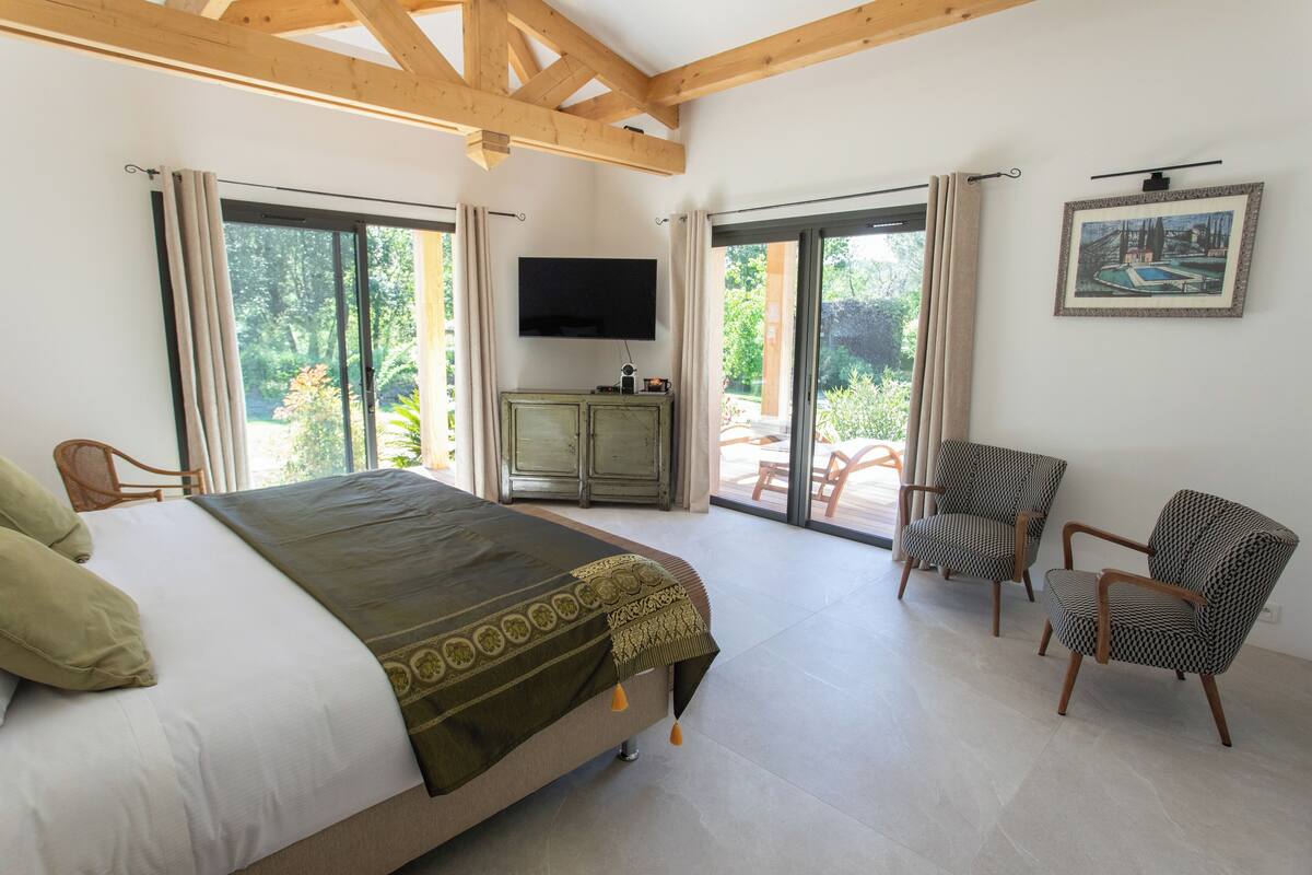 Un dormitorio con cama, televisor, dos sillas y un ventanal corredero que da a un espacio exterior.