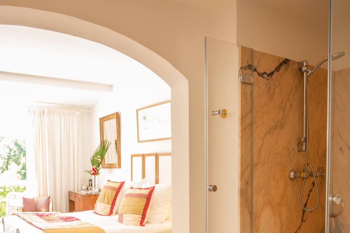 Habitación de hotel con ducha de mármol, cama con ropa de cama roja y beige, zona de estar con una silla y una mesa, y vistas al exterior.