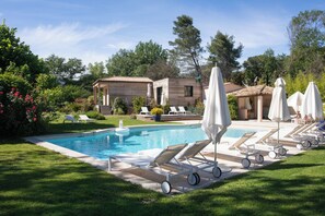 A swimming pool with lounge chairs and umbrellas, surrounded by greenery and a building with a tiled roof. - Le Verger Maelvi (Grimaud)