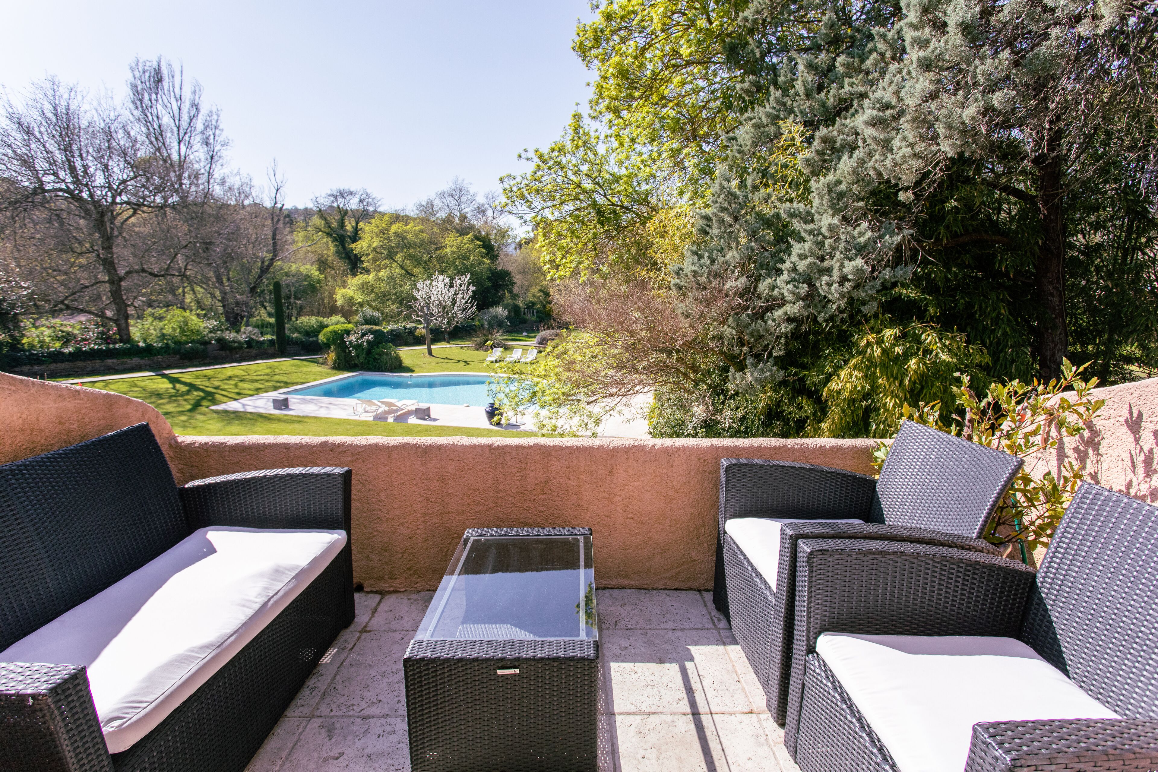 Un patio al aire libre con mobiliario de mimbre, una mesa con superficie de vidrio y vistas a una piscina y jardín.