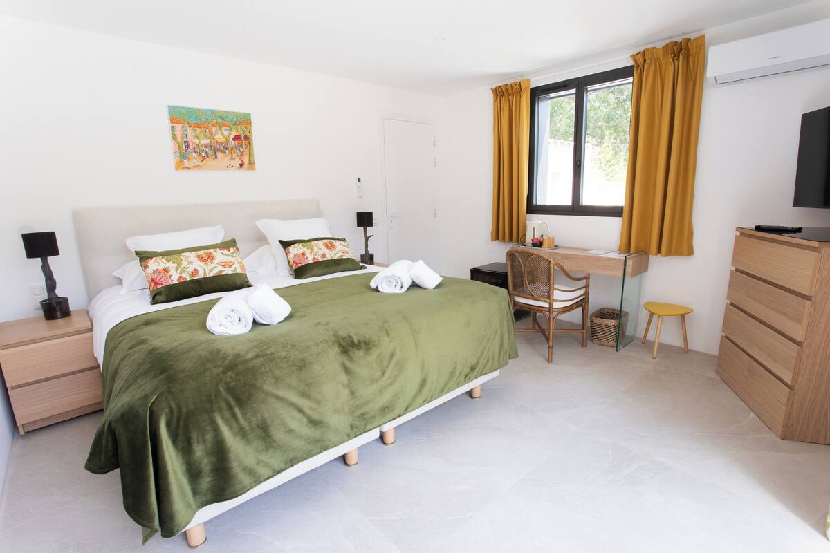 Un dormitorio ordenado con cama, mesitas de noche, escritorio, silla, televisor y ventana con cortinas.