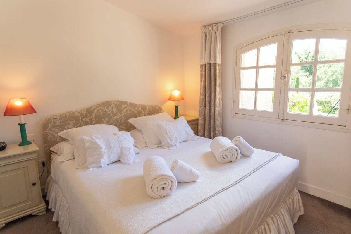 Una cama bien hecha con toallas enrolladas, dos lámparas de noche y una ventana con cortinas.