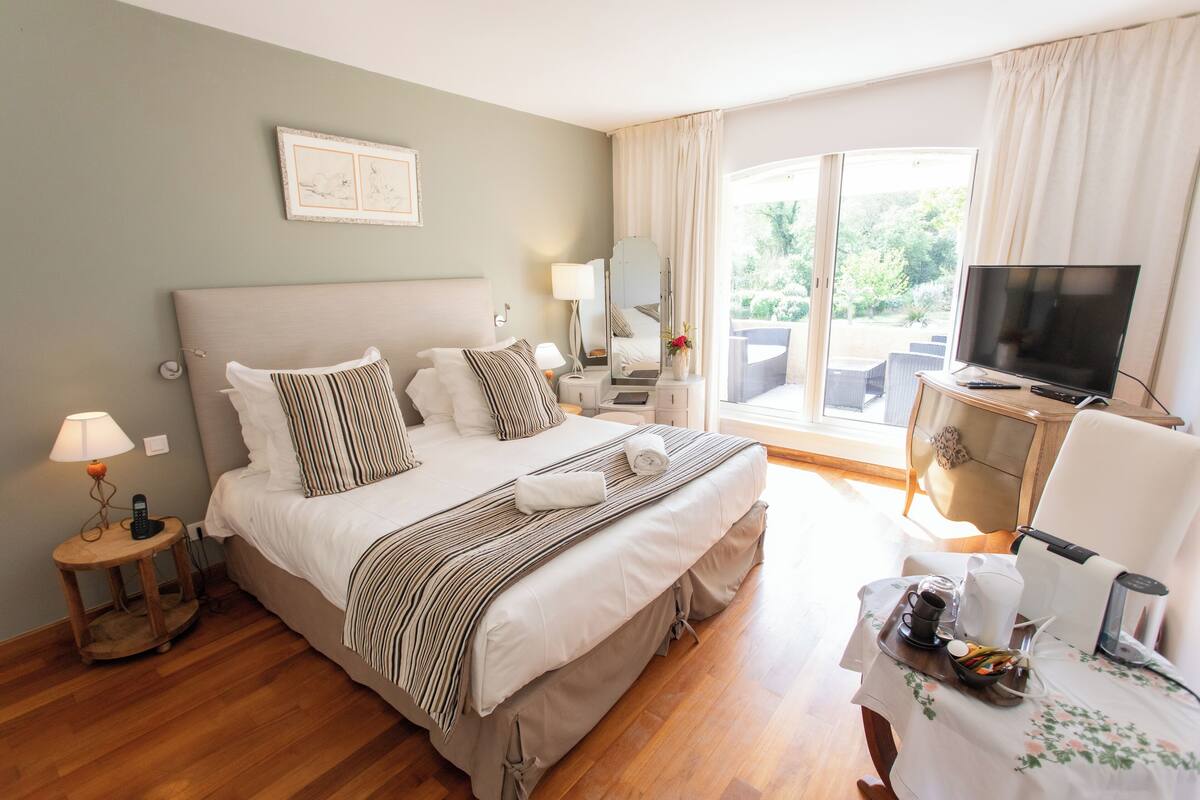 Habitación de hotel con una cama grande, un televisor, una mesita con bandeja y un balcón con mobiliario exterior.