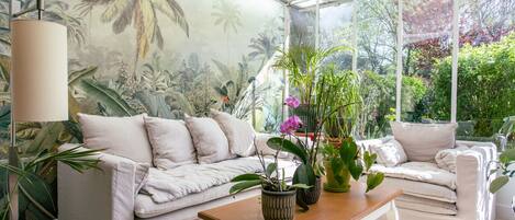 Um solário com um sofá branco, uma mesa de madeira e diversos vasos com plantas.