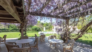 An outdoor patio with wicker furniture and a pergola covered in purple flowers. - Le Verger Maelvi (Grimaud)