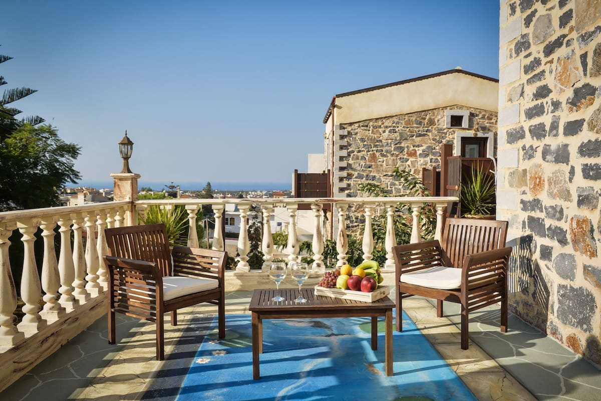 A balcony with a stone wall, two wooden chairs, a table with fruit and glasses, and a view of the sea.