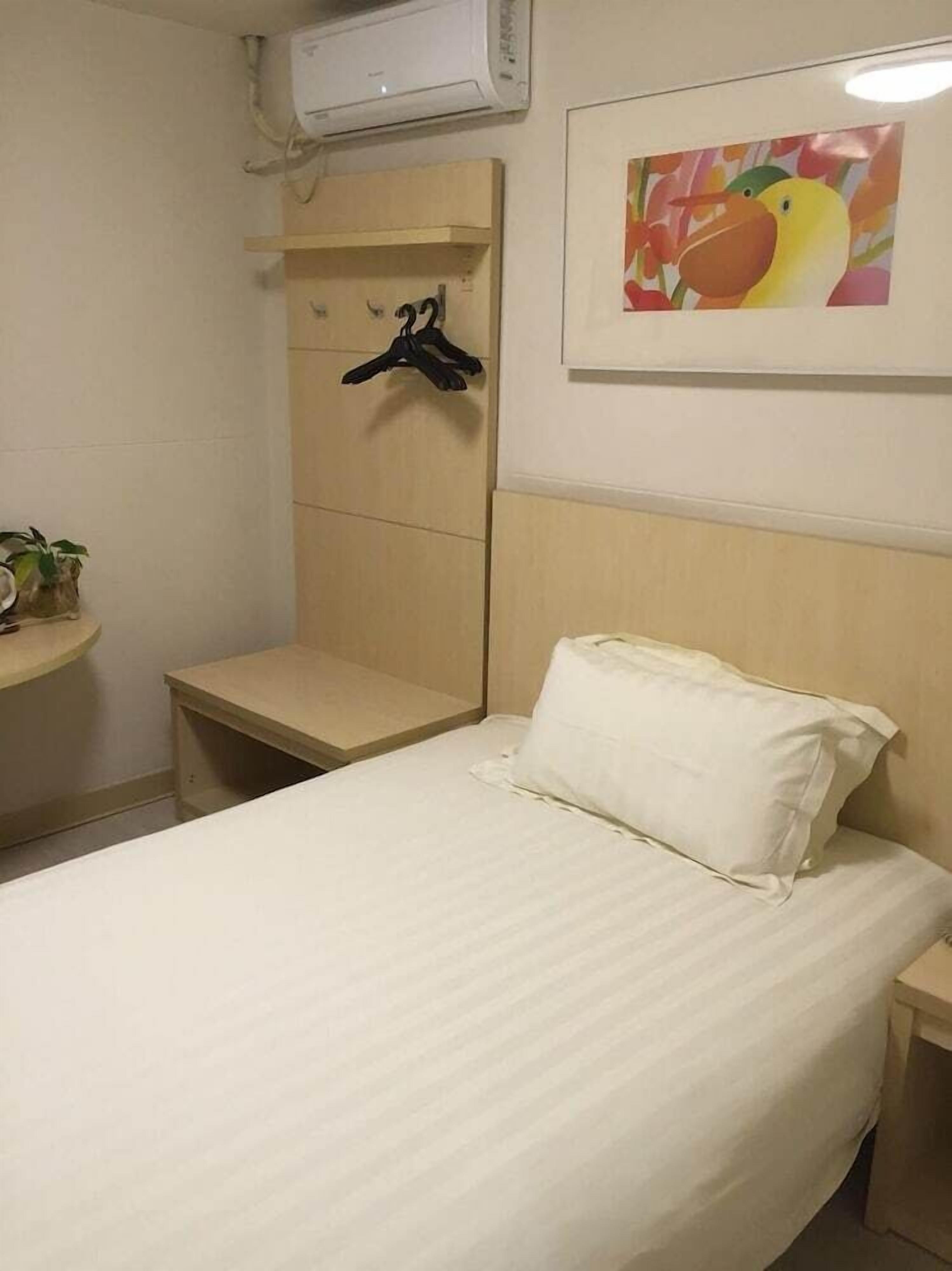 Una cama individual con un almohadón blanco y cabecera, una mesita de noche pequeña, un aire acondicionado instalado en la pared, un cuadro enmarcado y un perchero negro en un soporte de pared.