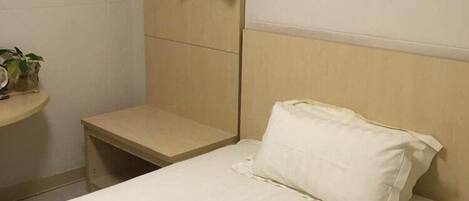 Una cama individual con un almohadón blanco y cabecera, una mesita de noche pequeña, un aire acondicionado instalado en la pared, un cuadro enmarcado y un perchero negro en un soporte de pared.