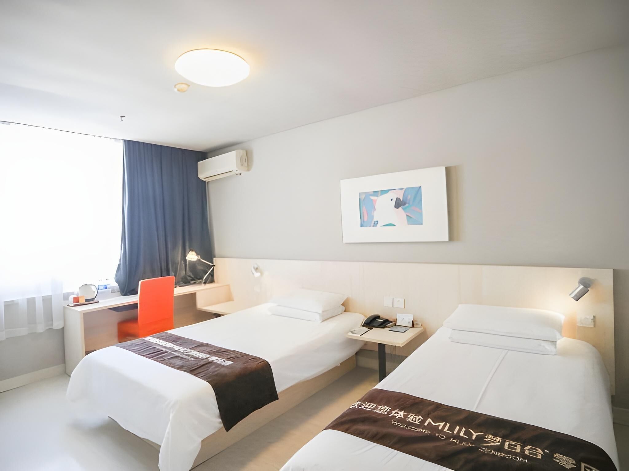 A hotel room with two beds, a desk, a chair, a desk lamp, a framed picture on the wall, and an air conditioning unit.