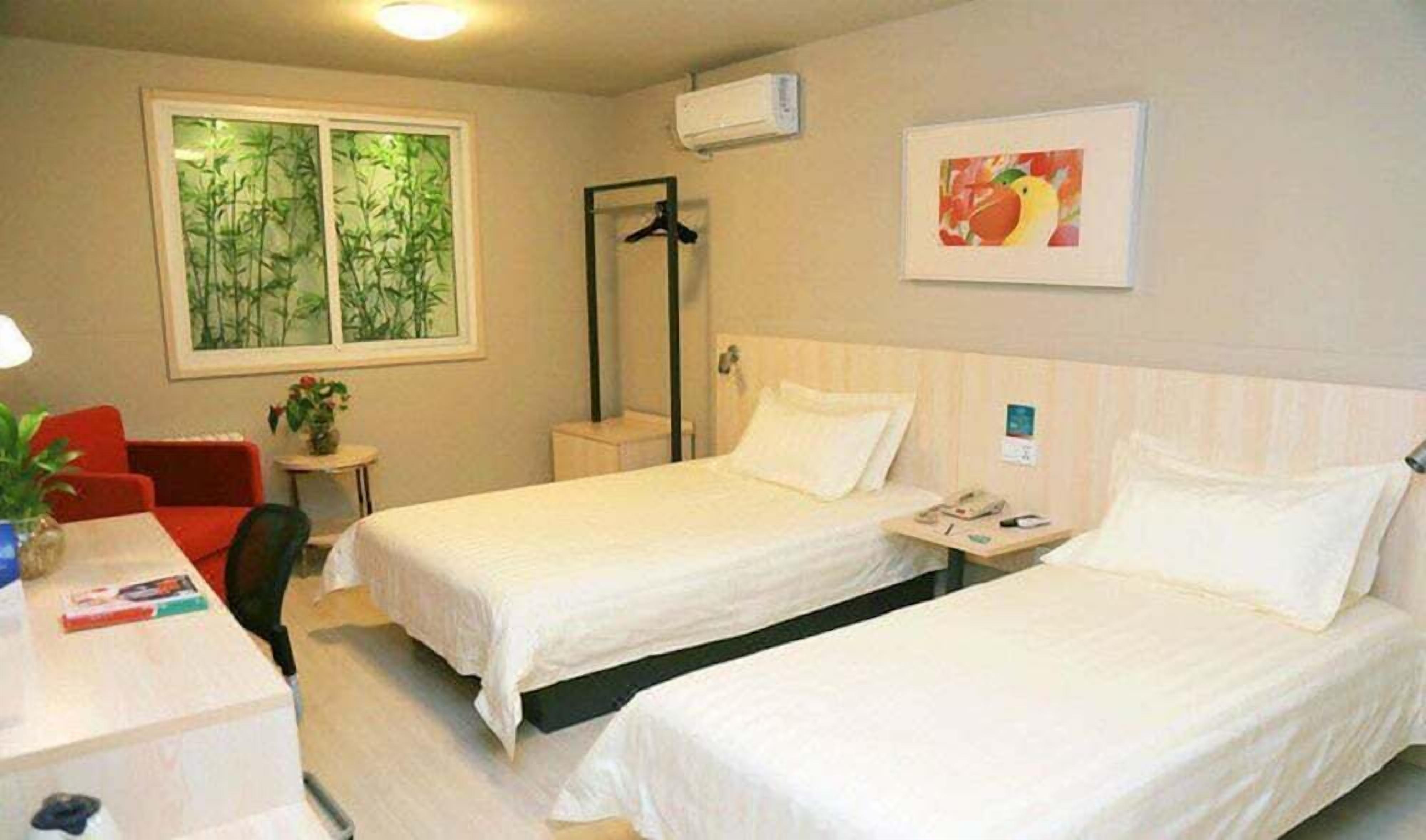A hotel room with two beds, a desk with a chair, a small table with a plant, a window with a bamboo view, and a framed picture on the wall.