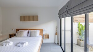 A modern hotel room with a large bed, a wooden headboard, a bedside table with a lamp, a small wooden shelf, and a view of the outside through large windows. - Studio365 Apartment (Chiang Mai)