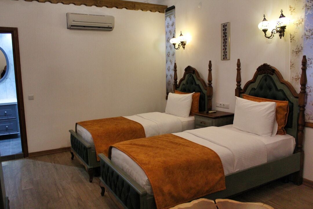A room with two beds, each with a wooden headboard, white bedding, and a brown throw blanket. There is a nightstand with a lamp between the beds, and a wall-mounted air conditioner above the left bed.