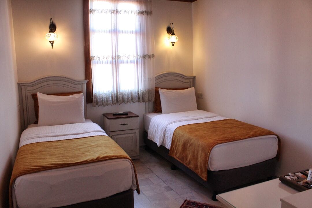 Een hotelkamer met twee eenpersoonsbedden, elk met een houten hoofdbord, witte beddengoed en een bruine plaid. Er zijn nachtkastjes met lampen en een raam met dunne gordijnen.