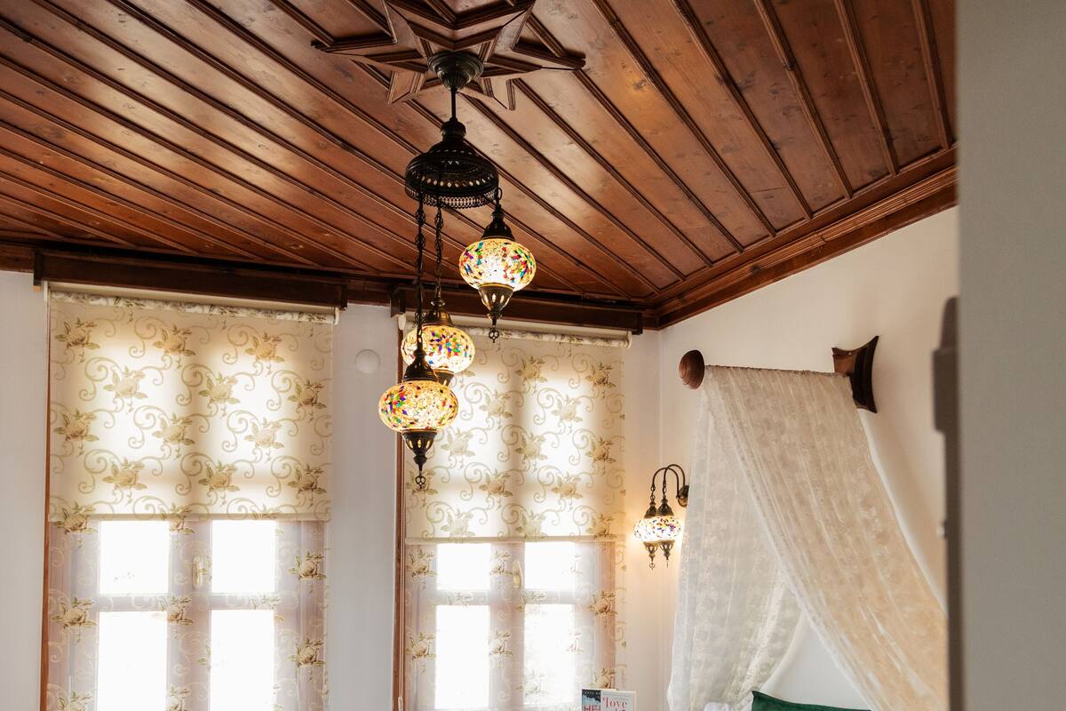 A room with a wooden ceiling, a bed with a green bedspread, two windows with floral curtains, a nightstand with a cup, and a ceiling fan.