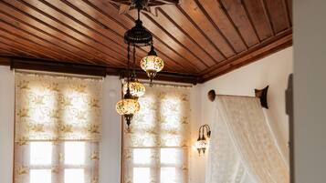 A room with a wooden ceiling, a bed with a green bedspread, two windows with floral curtains, a nightstand with a cup, and a ceiling fan.