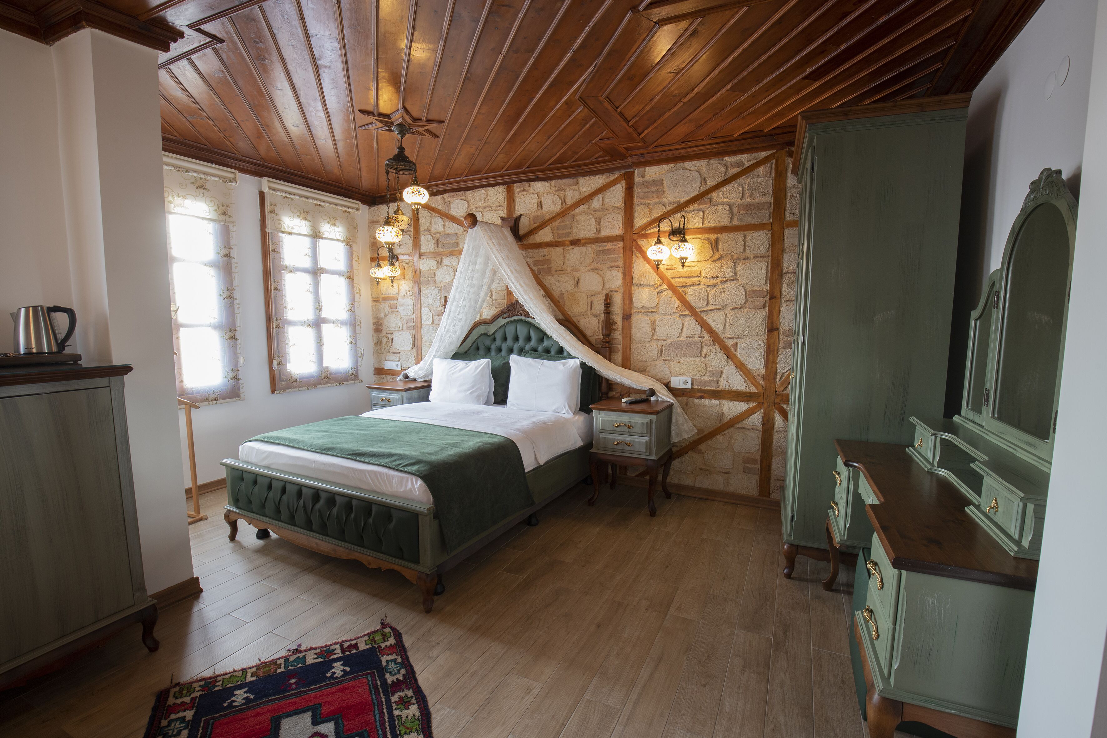 A bedroom with a four-poster bed, a stone wall, wooden beams, a green upholstered headboard, a nightstand, a bedside table, a dresser, and a mirror.