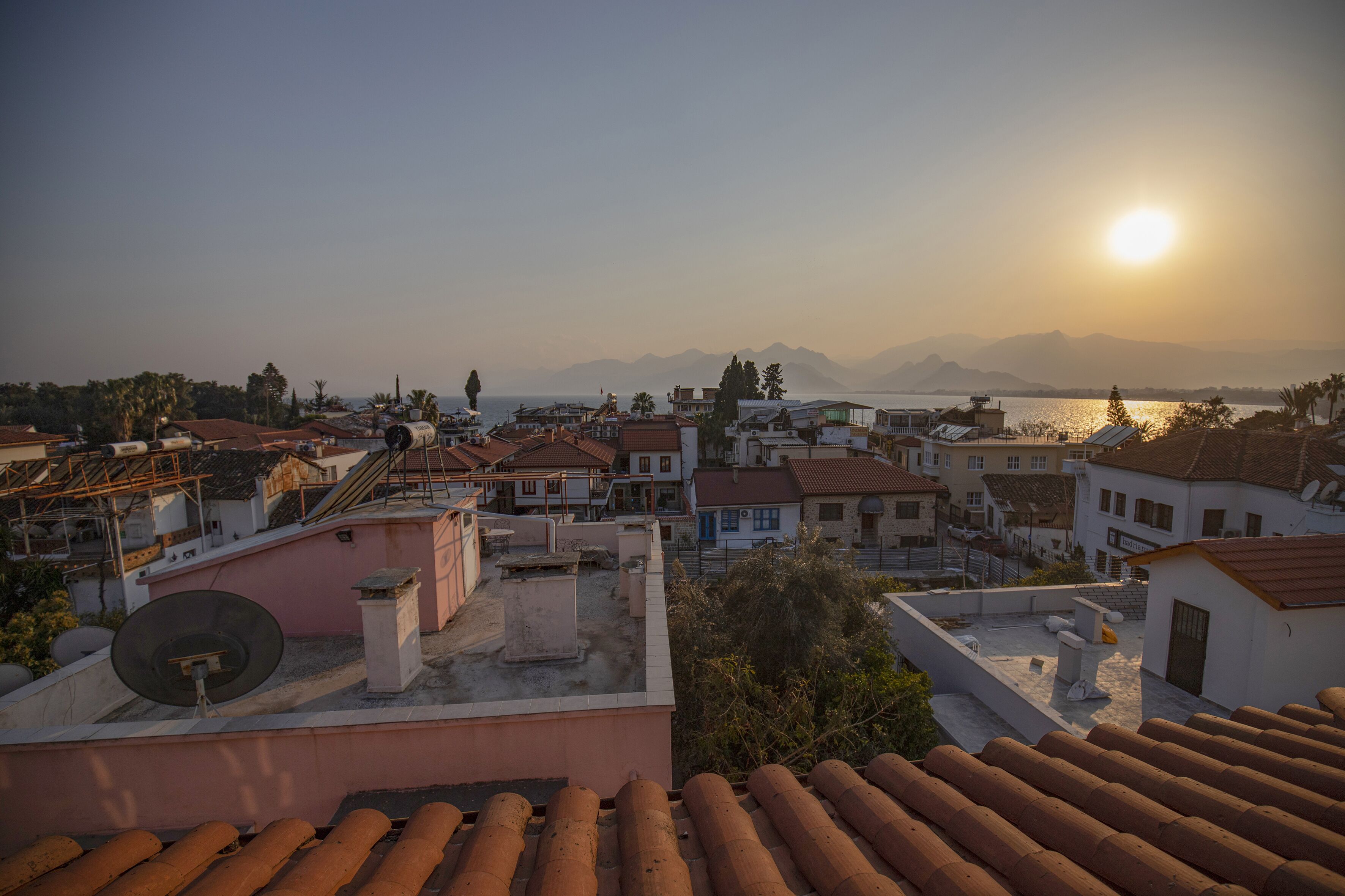 A coastal town with terracotta-roofed buildings, a clear sky, and the sun setting over the sea.