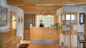 A wooden reception desk with a person standing behind it, a large window with multiple panes, and a clock on the wall. - Hotel Garni Belvedere, das BIO Hotel & SuiteHotel am Ederse (Waldeck)