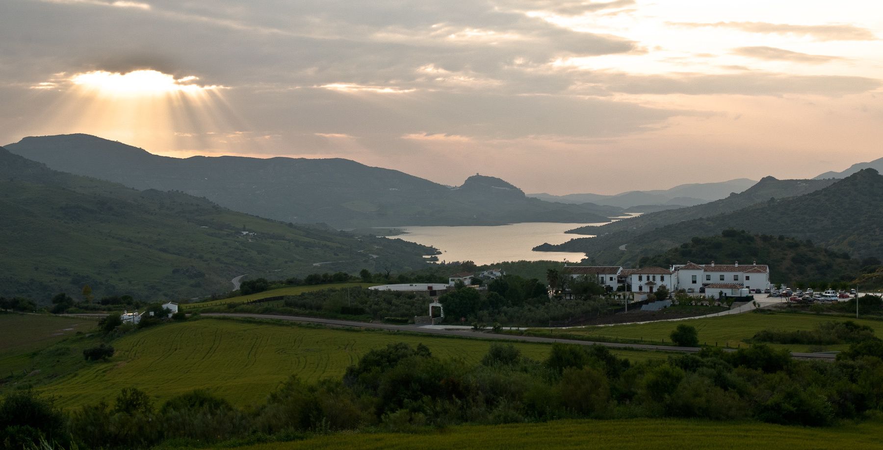 Paisatge rural amb un llac, turons ondulats i un edifici blanc amb diverses finestres i teulada vermella.