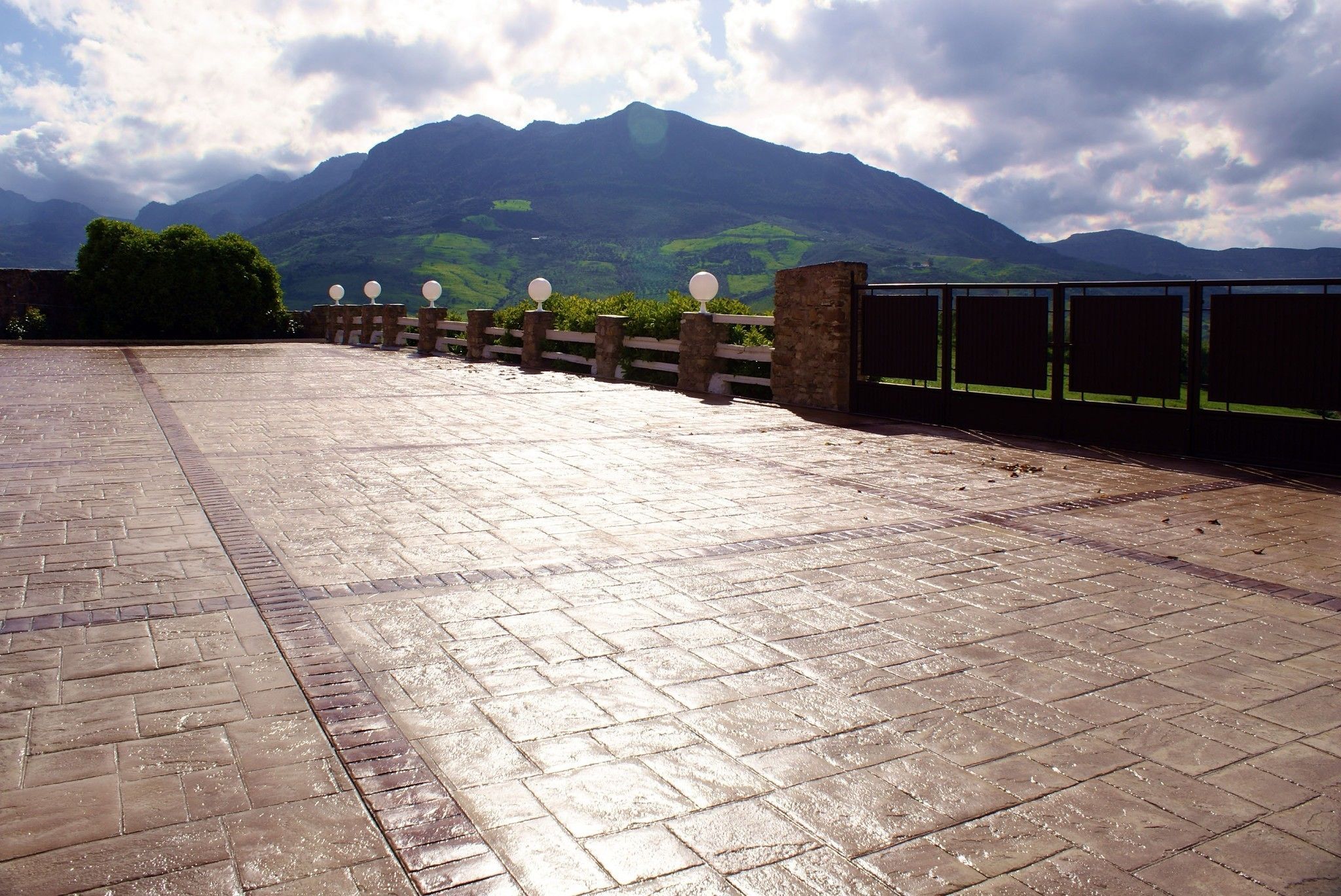 Un chemin pavé, bordé d’une rambarde en pierre et de lampadaires, qui mène à un point de vue sur la montagne.