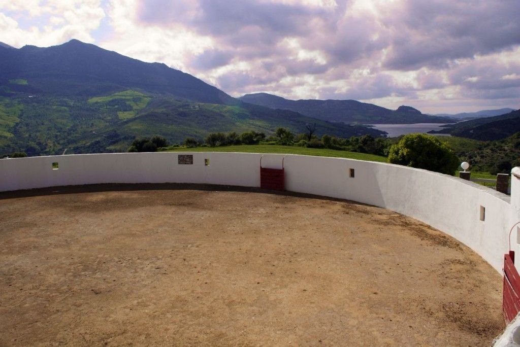 A circular outdoor area with a dirt floor, surrounded by a white wall with a red door, overlooking a mountainous landscape.