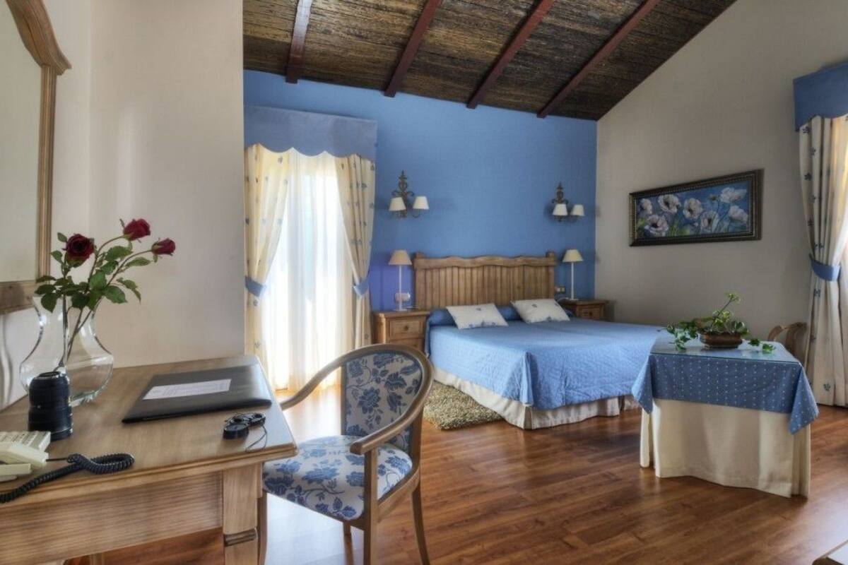 A bedroom with a wooden desk, a chair, a bed with a blue bedspread, a nightstand, a lamp, a painting on the wall, and a window with curtains.