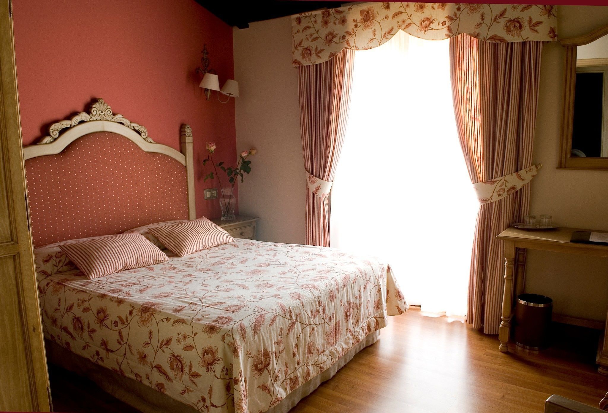 Un dormitorio con cubrecama floral, cabecera de madera, mesita de noche con lámpara y ventana con cortinas.