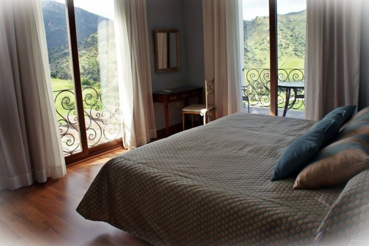 A bedroom with a large bed, a chair, a desk, and a balcony with a view of the countryside.
