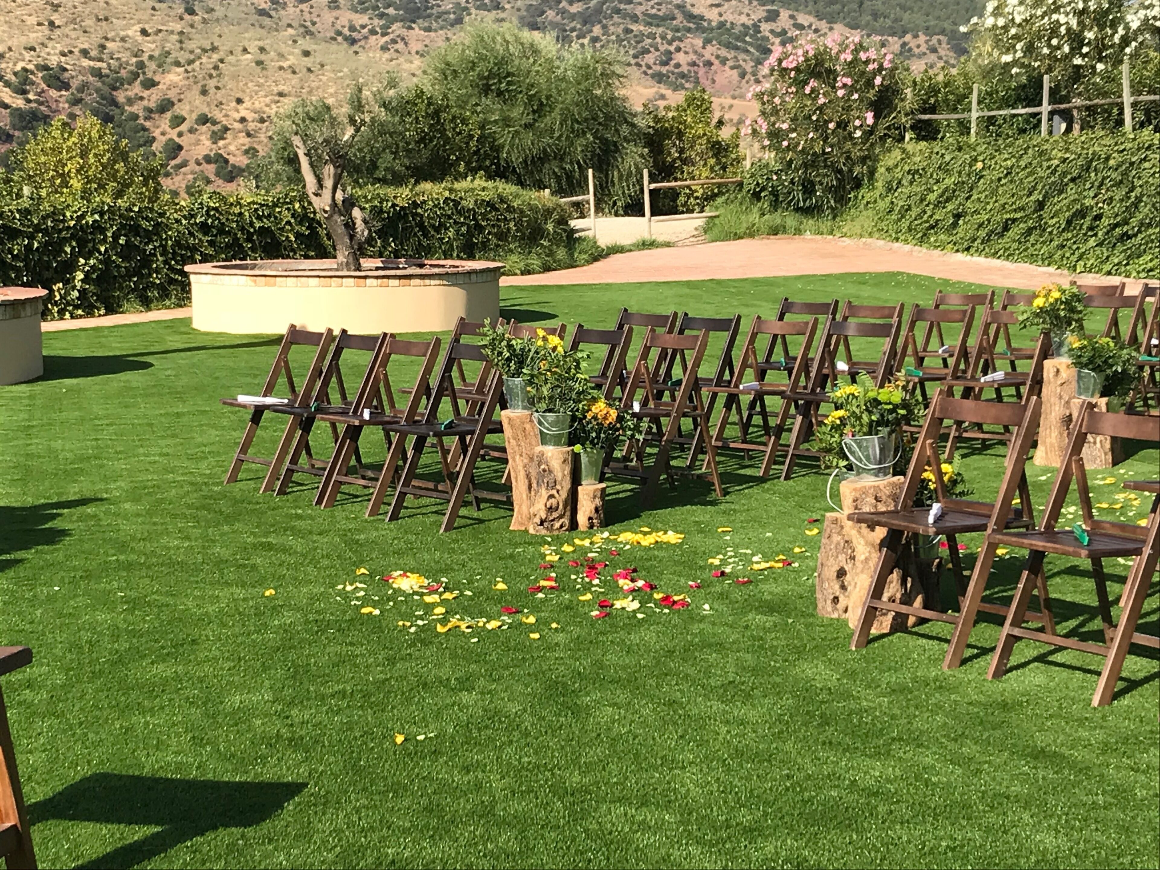 Montaje para una ceremonia de boda con sillas de madera dispuestas en filas sobre un césped, rodeado de follaje y colinas al fondo.