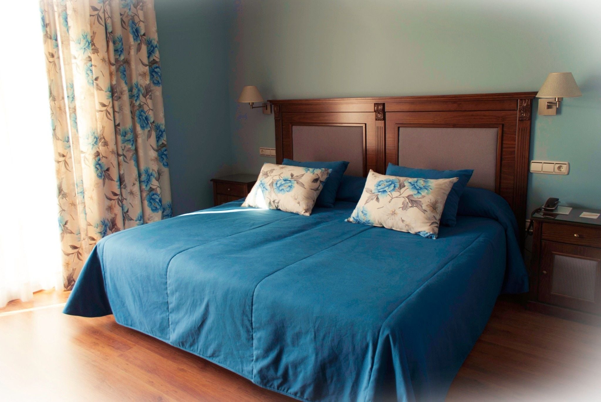 A neatly made bed with blue bedding and floral pillows, a wooden headboard, and a nightstand with a lamp.