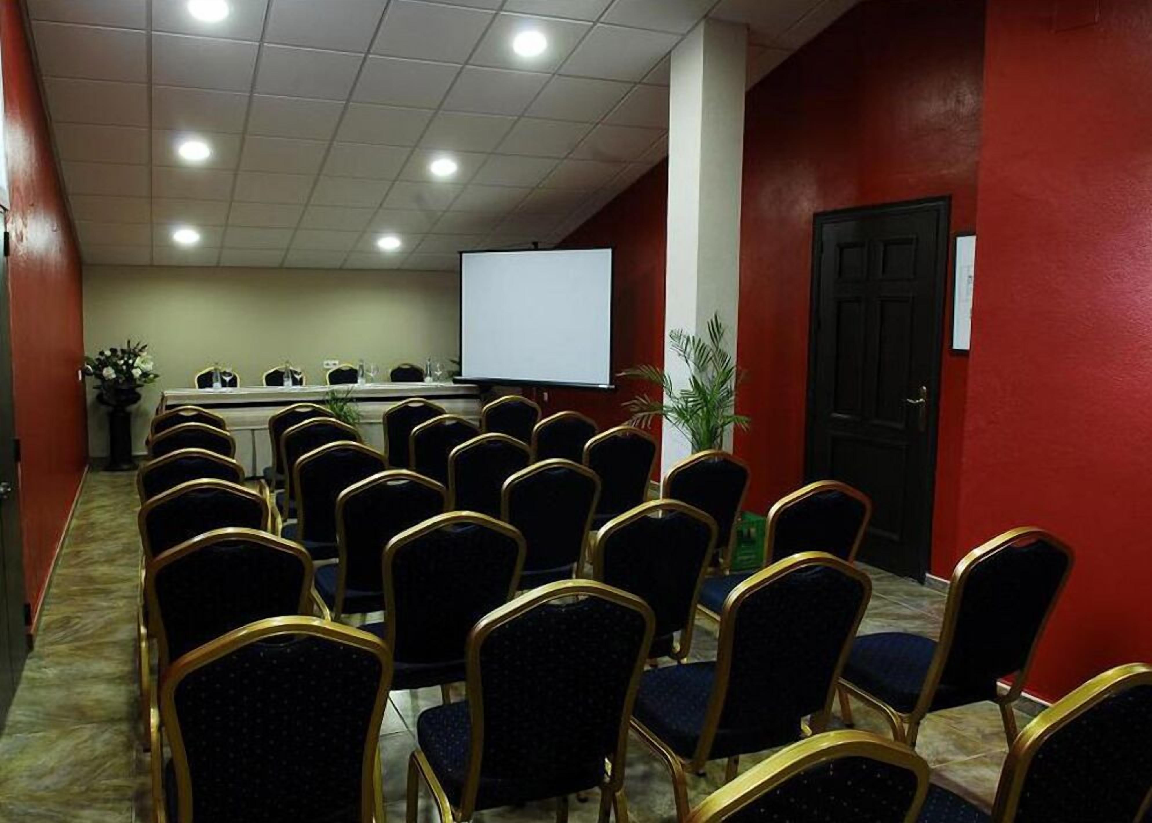 Un salón de conferencias con filas de sillas orientadas hacia una pizarra, un atril y una pantalla de proyector.