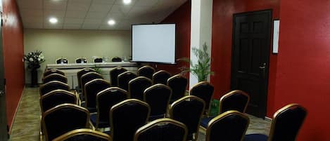 Un salón de conferencias con filas de sillas orientadas hacia una pizarra, un atril y una pantalla de proyector.