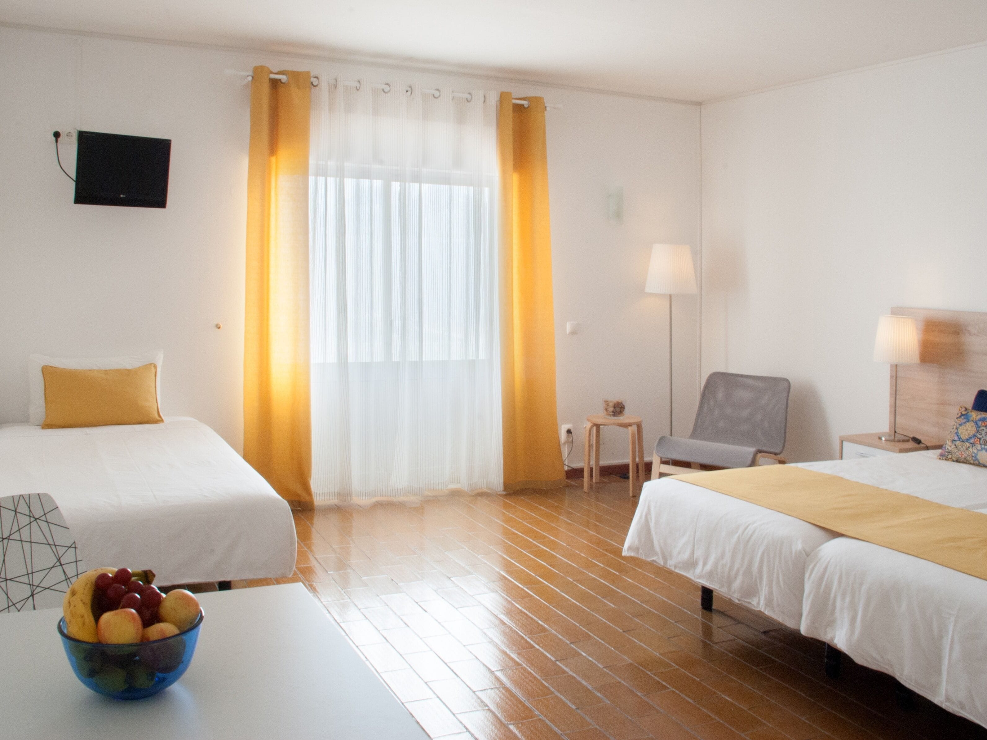 Une chambre d’hôtel avec deux lits, une petite table sur laquelle se trouve un panier de fruits, une chaise, une lampe et un téléviseur fixé au mur.