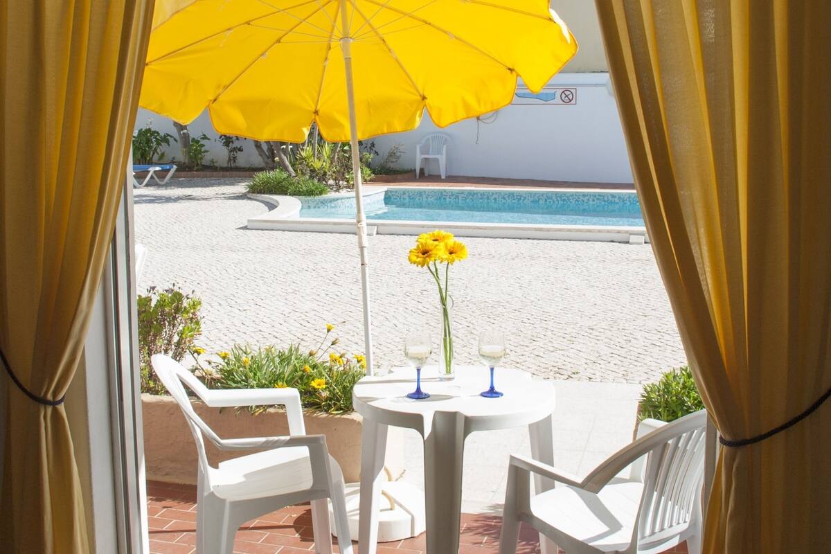 A terrace with a yellow umbrella, white chairs, a table with two vases of flowers, and a pool in the background.